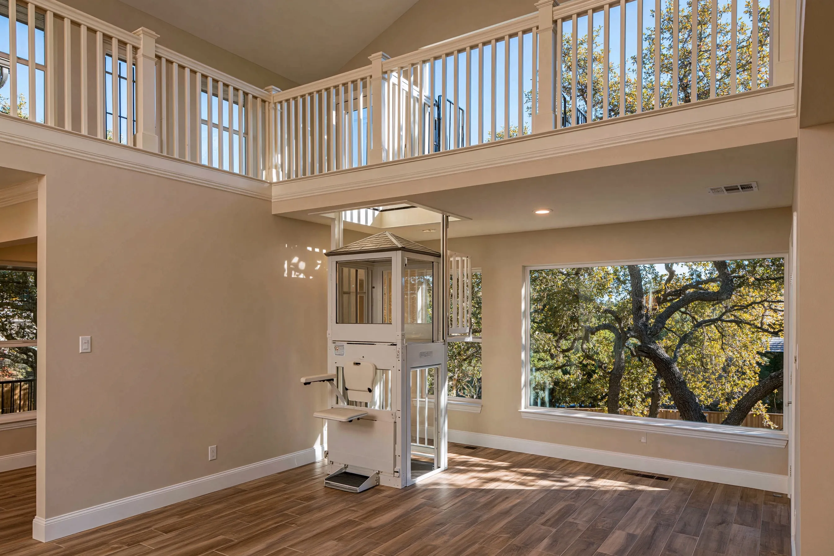 Compact white through-floor lift with a fold-down seat in the corner of a two-story family room, below a framed opening in the loft floor and the loft railing
