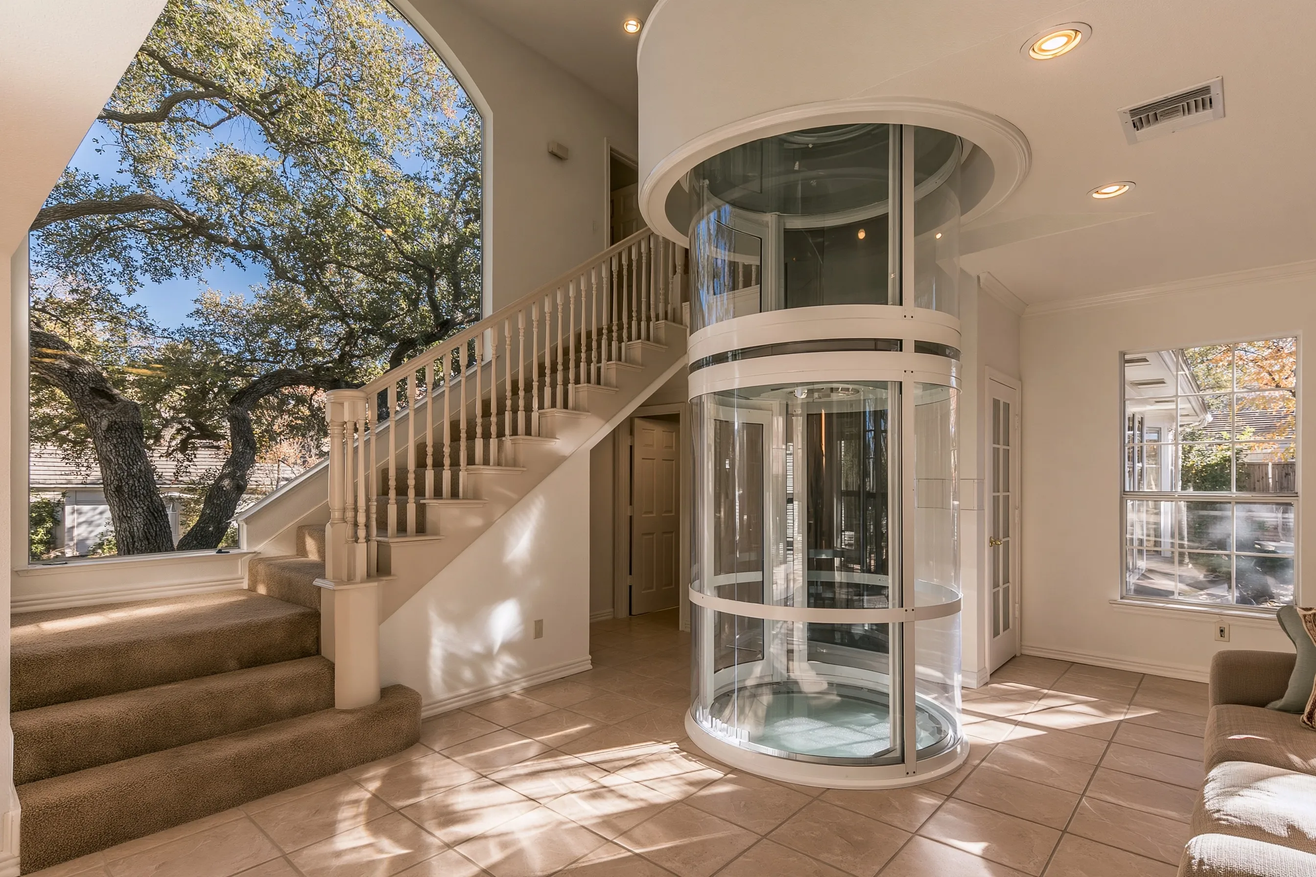 Wide round see-through elevator standing on the tile floor beside a carpeted stair, rising into a round opening in the ceiling